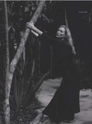 Kate Moss wearing a long black dress while leaning against a tree trunk in an outdoor wooded setting in black and white.