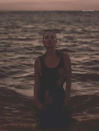 Kate Moss stands waist-deep in the dark ocean water at dusk, wearing a sleeveless black top.