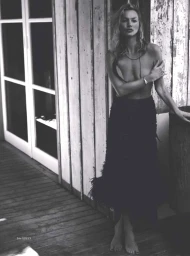 Monochrome portrait of Kate Moss posing topless in a textured black skirt against a weathered wood paneled wall.