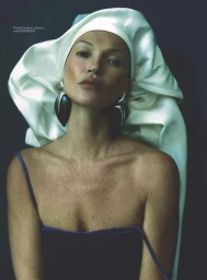 Kate Moss wearing a voluminous white satin headdress, oversized statement earrings, and a navy velvet top against a dark backdrop.