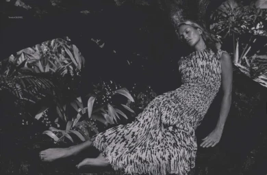 Monochrome portrait of Kate Moss reclining barefoot against a tree trunk wearing a textured sleeveless patterned dress.