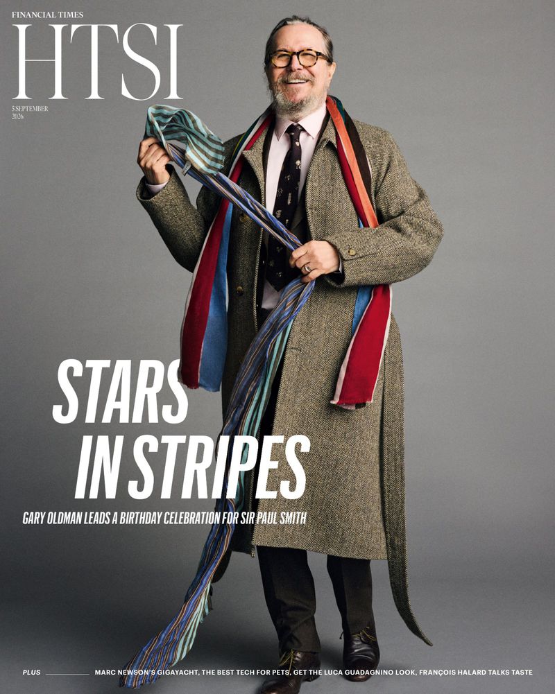 Actor Gary Oldman smiling in a tweed coat and eyeglasses holding a long colorful striped scarf against a grey backdrop.