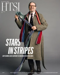 Actor Gary Oldman smiling in a tweed coat and eyeglasses holding a long colorful striped scarf against a grey backdrop.