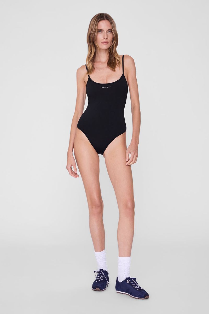 Model wearing a black spaghetti-strap bodysuit, white socks, and navy sneakers against a seamless light grey studio backdrop.