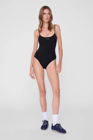 Model wearing a black spaghetti-strap bodysuit, white socks, and navy sneakers against a seamless light grey studio backdrop.
