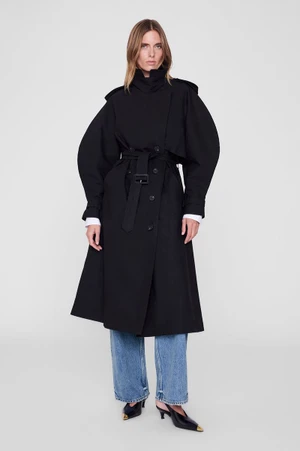 Model wearing an oversized black belted trench coat and blue denim jeans against a seamless light studio background.