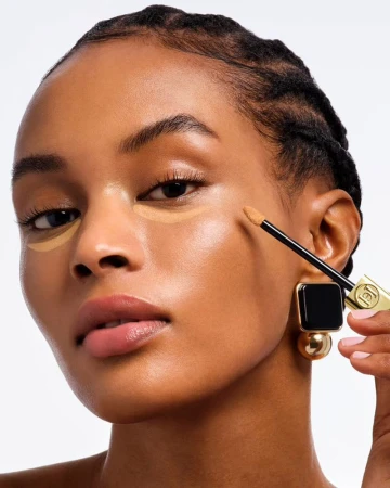 Model Bineta Loupdy applying under-eye concealer with a wand applicator against a bright white seamless studio background.