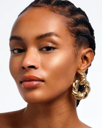 Close-up beauty portrait of Bineta Loupdy with glowing skin wearing a chunky gold twisted hoop earring against a white background.