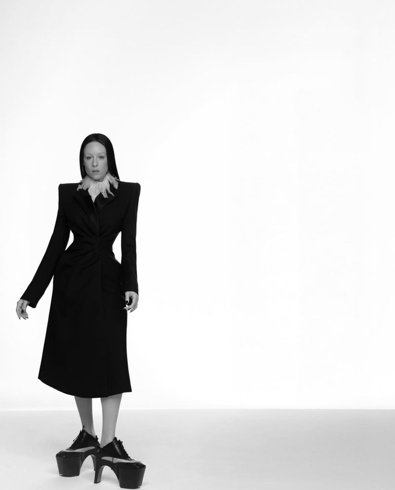 Woman in black dress and high platform shoes stands in a minimalist white setting, posing for a fashion-forward, monochromatic photo.