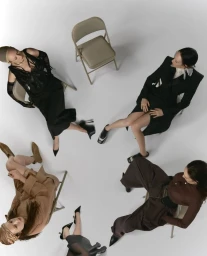 Four seated women surrounding an empty folding chair against a white studio background in an editorial fashion shot by Arseny Jabiev.