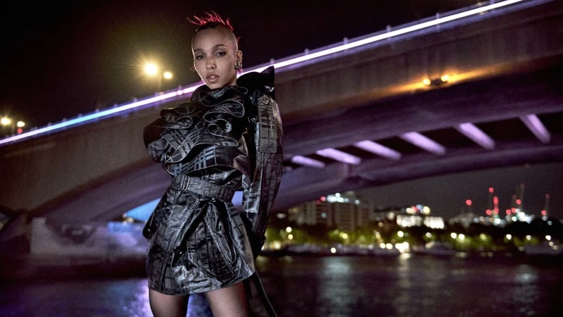 FKA Twigs wearing a dark patterned trench coat standing outdoors at night beneath a illuminated bridge for Burberry