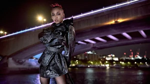 FKA Twigs wearing a dark patterned trench coat standing outdoors at night beneath a illuminated bridge for Burberry