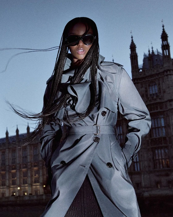FKA Twigs wearing a grey belted trench coat and dark sunglasses against a gothic building exterior at dusk.