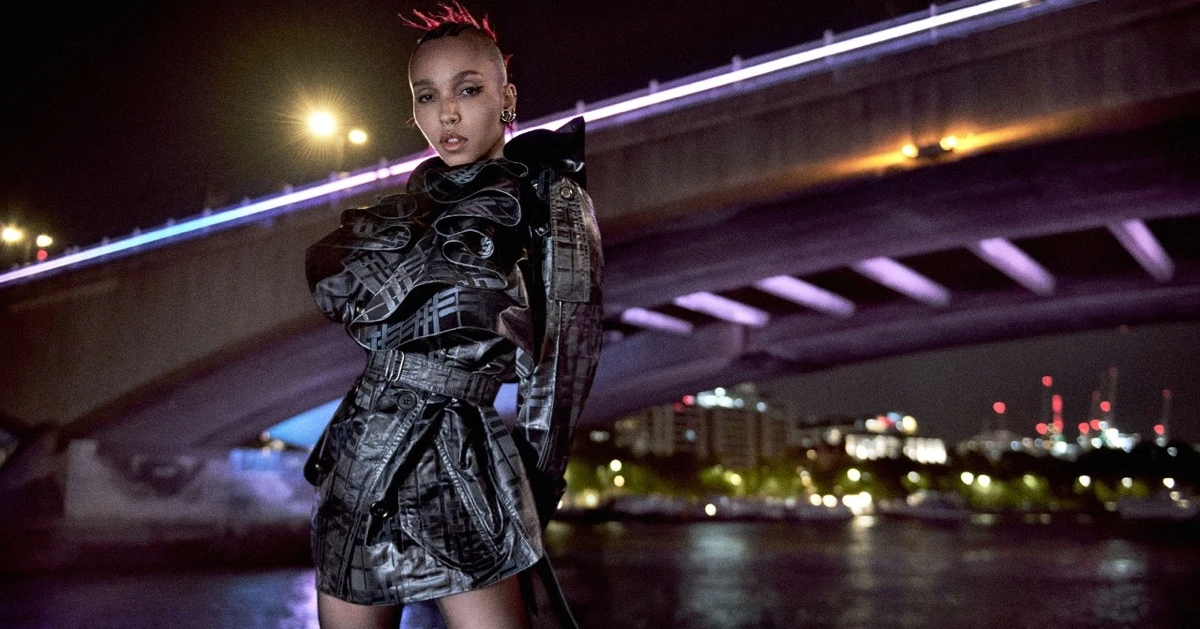 FKA Twigs in a dark patterned belted coat posing beneath an illuminated bridge at night for Burberry Winter 2026