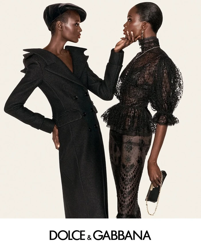 Two black female models in black designer clothing against an off-white studio background for Dolce & Gabbana.