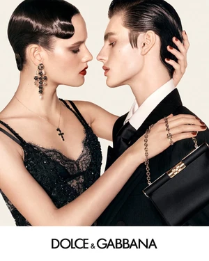Two models facing each other closely wearing black formal attire against a light background with Dolce Gabbana branding below