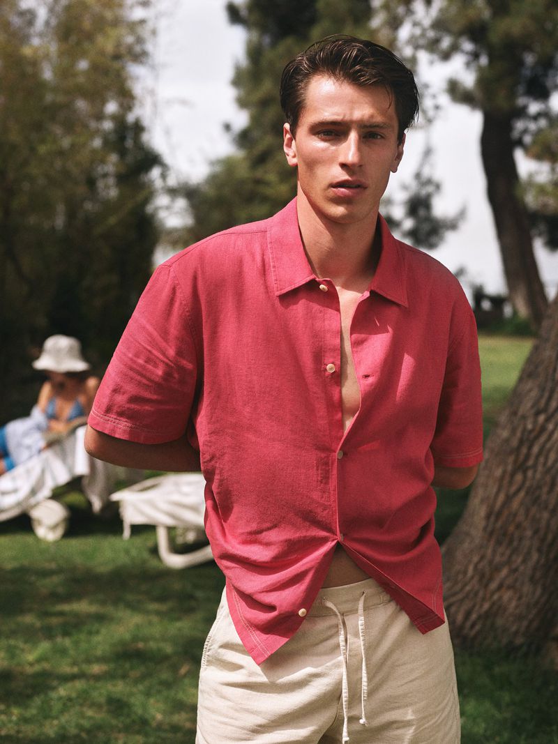 Portrait of a young man wearing a pink linen shirt and beige shorts, standing outside on a sunny day with trees and lawn in the background.