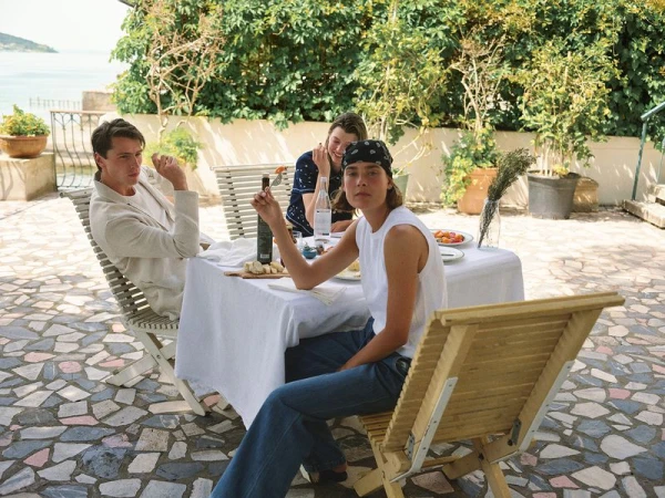 Three people sit at a table with food and drinks outdoors on a patio with trees and a water view in the background on a sunny day.