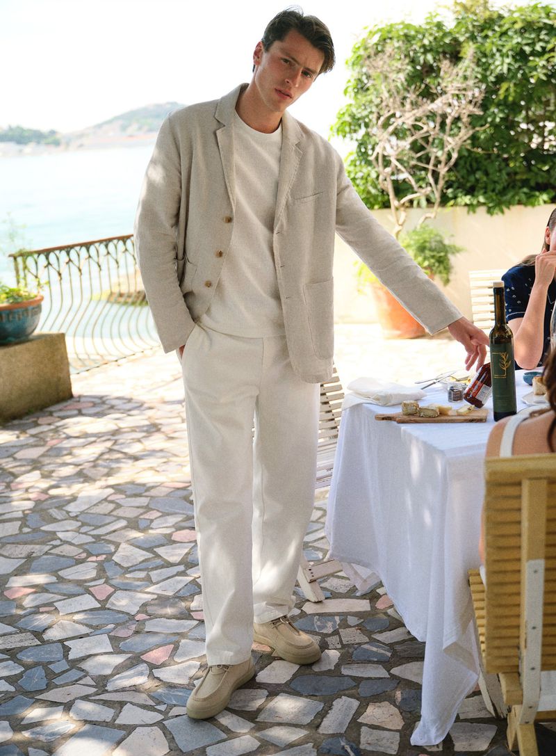 A stylish man in a light suit stands on a stone patio next to a table set for lunch by the sea on a bright, sunny day.