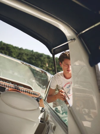 A man inside a white boat, with green trees visible in the background under a bright sky, enjoying a sunny day on the water.