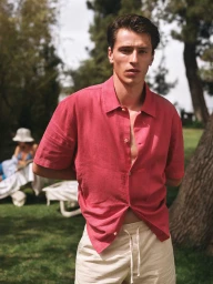 A man wearing a pink short-sleeve button-down shirt and light-colored drawstring trousers standing outdoors on a sunny lawn.