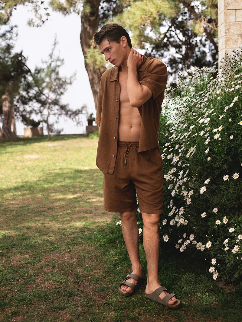 Model wearing brown linen shirt and shorts standing outdoors near white wildflowers and trees for Mavi