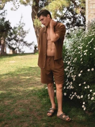 Model wearing brown linen shirt and shorts standing outdoors near white wildflowers and trees for Mavi