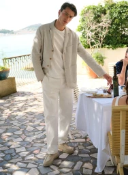 Man wearing a beige linen blazer and white trousers standing outdoors by a dining table near the sea.