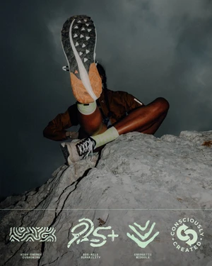 Low-angle shot of a person on a rocky crag holding up a trail running shoe to display the sole against a dark overcast sky.