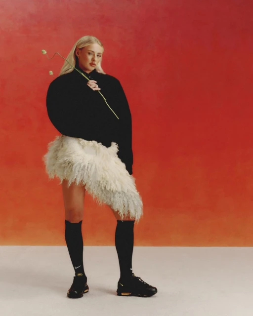 Iggy Azalea poses in black knitwear and fluffy skirt against an orange studio backdrop, holding a flower, and wearing black Nike shoes.