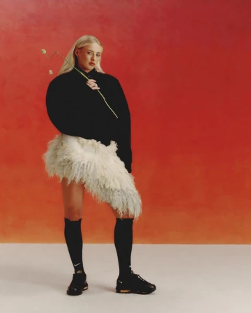 Iggy Azalea poses in black knitwear and fluffy skirt against an orange studio backdrop, holding a flower, and wearing black Nike shoes.