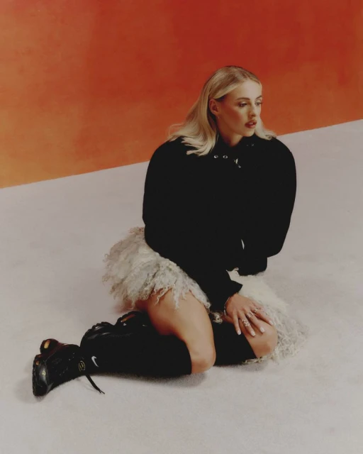 Blonde woman in black sweatshirt and tulle skirt kneeling on a white surface with orange background in a studio shot.