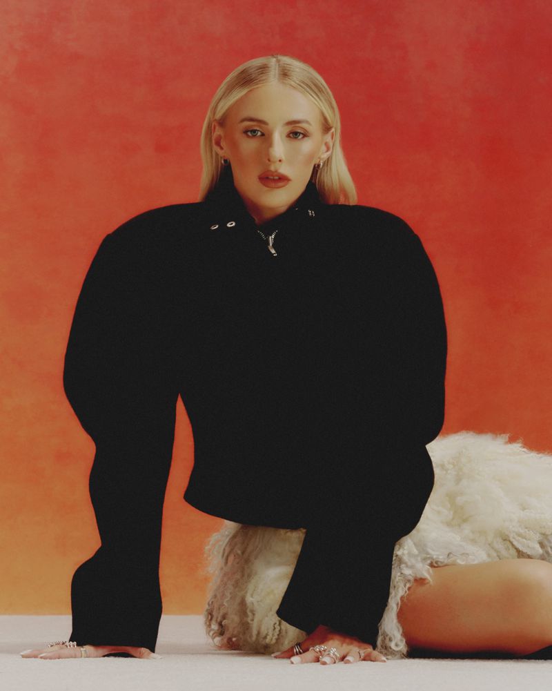 Young woman in black crop top and skirt reclines against an orange background in a studio setting, modelling rings and jewelry.