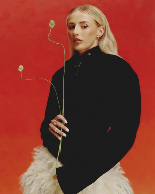Portrait of a woman with light hair in a black coat, holding a plant against a red background, showcasing fashion and style.