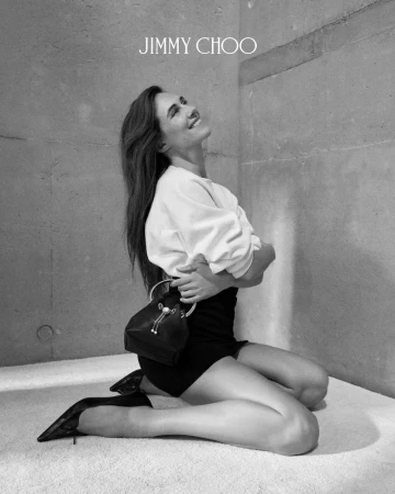 Devon Lee Carlson kneeling in a white top, black skirt, and pointed heels holding a small black handbag against a concrete wall.