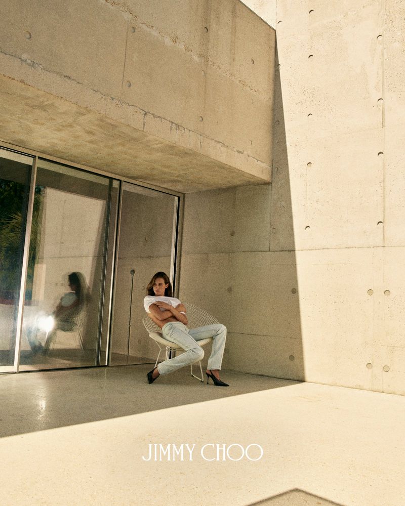 Devon Lee Carlson sits in a wire chair wearing a white top, light blue jeans, and black slingback heels against a concrete wall.