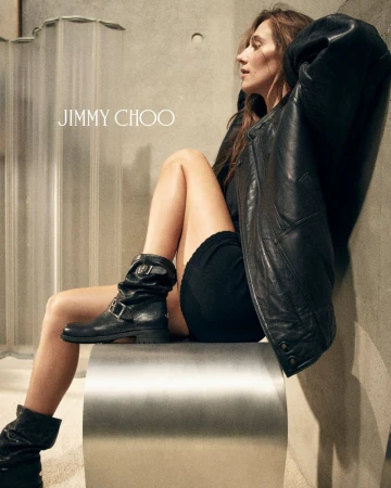 Devon Lee Carlson wearing a black leather jacket and slouchy buckle boots while seated on a curved metal stool against a concrete wall.