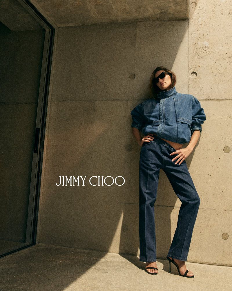 Devon Lee Carlson poses in a denim jacket, matching jeans, sunglasses, and black heels against a concrete wall for Jimmy Choo.