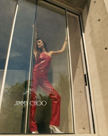 Devon Lee Carlson wearing a red one-shoulder top and matching trousers behind a large glass door in a Jimmy Choo campaign.
