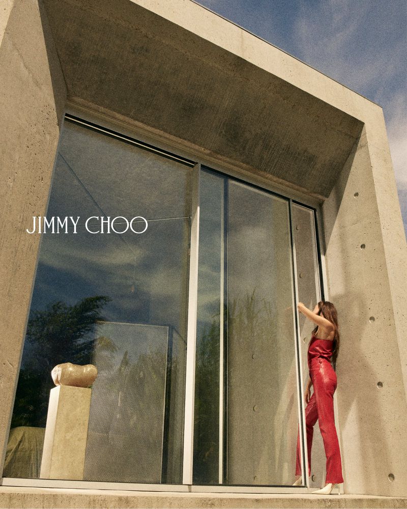 Devon Lee Carlson wearing a red jumpsuit standing by large glass sliding doors in front of a concrete building.