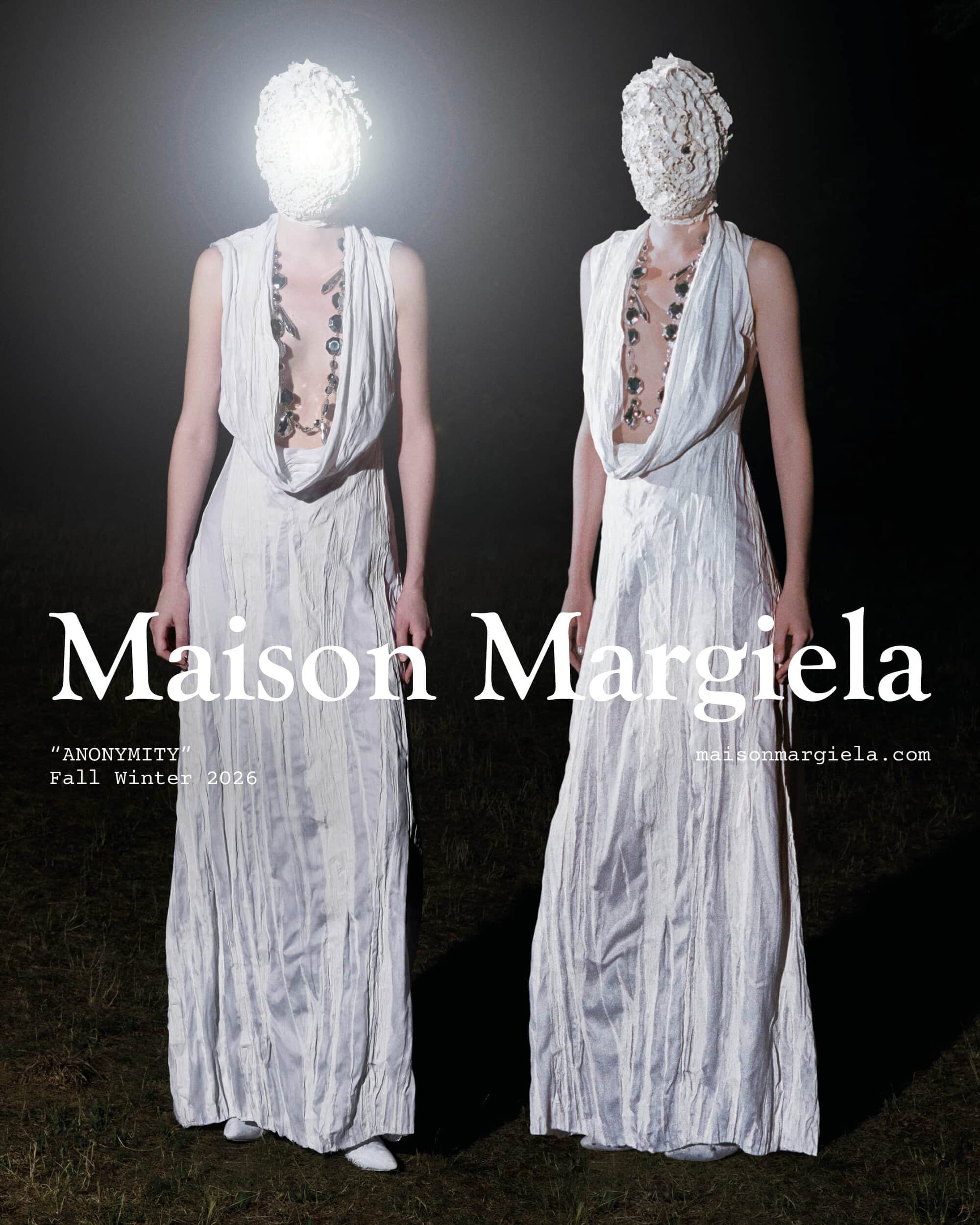 Two figures in textured white face coverings and draped white gowns standing against a dark outdoor night background.
