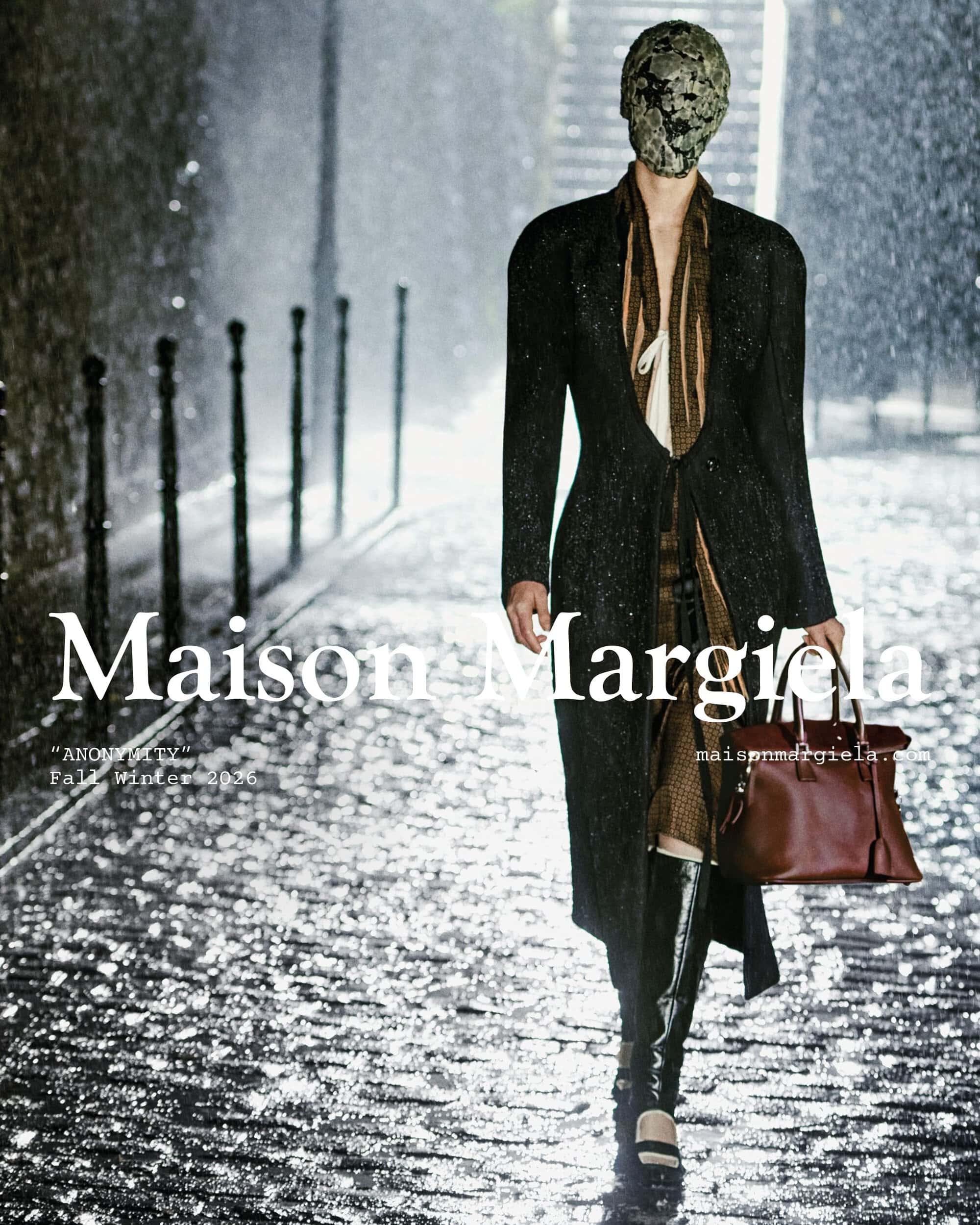 Model wearing a full face mask, long black tailored coat, and high boots carrying a brown handbag on a wet cobblestone street in rain.