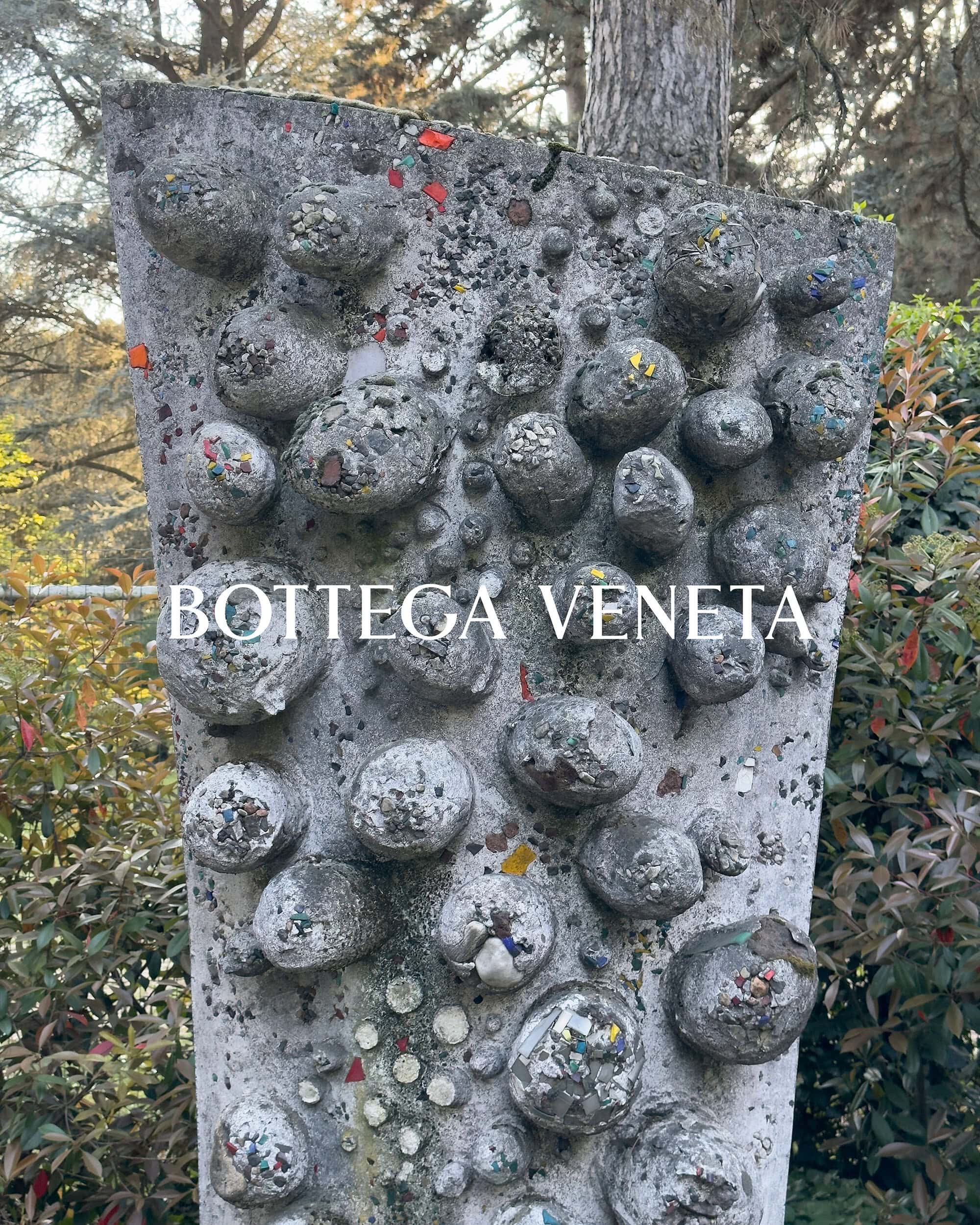 Stone sculpture embedded with rounded rocks and mosaic pieces in an outdoor garden with Bottega Veneta text overlay.