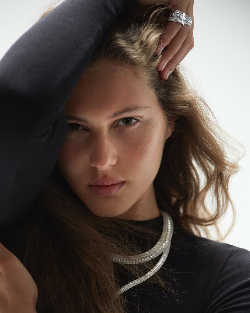 Close-up portrait of an adult female wearing a double-strand diamond necklace and a diamond ring on her finger against a white background.