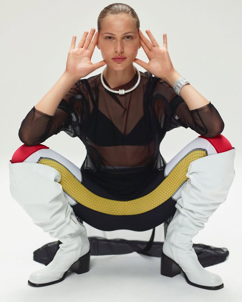 Woman in futuristic-style athletic wear poses in photo studio, showcasing a sheer black top, form-fitting leggings, and chunky white boots.