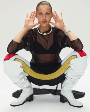 Woman in futuristic-style athletic wear poses in photo studio, showcasing a sheer black top, form-fitting leggings, and chunky white boots.