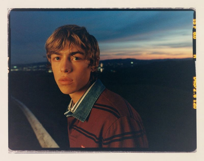 Portrait of a young man wearing a burgundy striped shirt with a denim collar against a dusk sky with distant city lights.