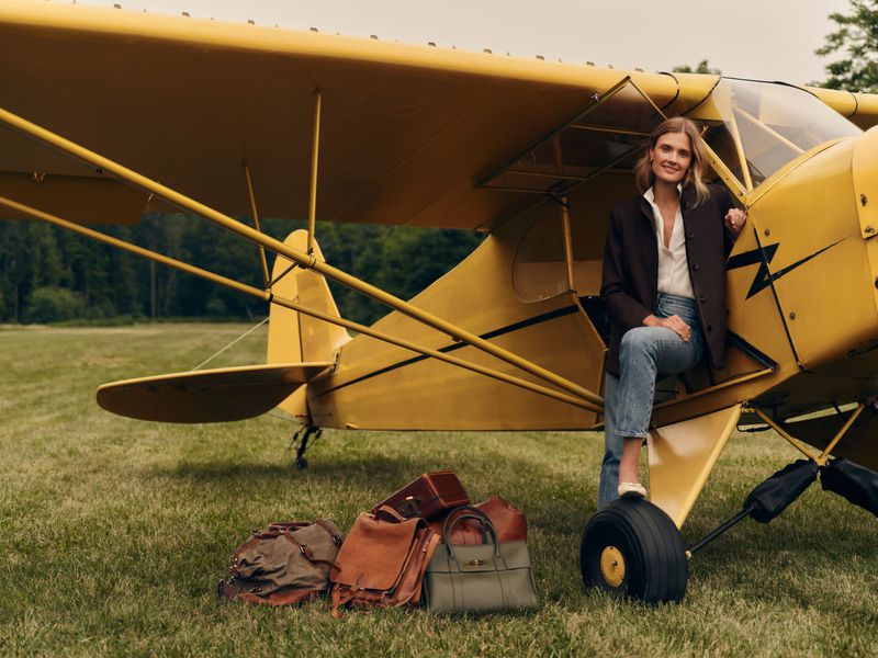 Model Constance Jablonski sits in the doorway of a yellow propeller plane with travel bags on a grassy field for Tuckernuck.