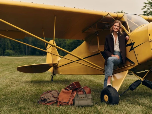 Model Constance Jablonski sits in the doorway of a yellow propeller plane with travel bags on a grassy field for Tuckernuck.