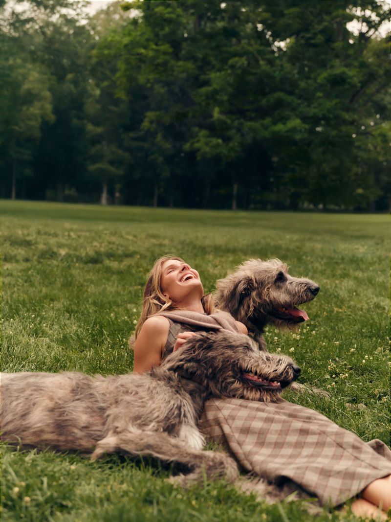 Constance Jablonski laughing while lying on a grassy meadow surrounded by two large Irish wolfhounds.
